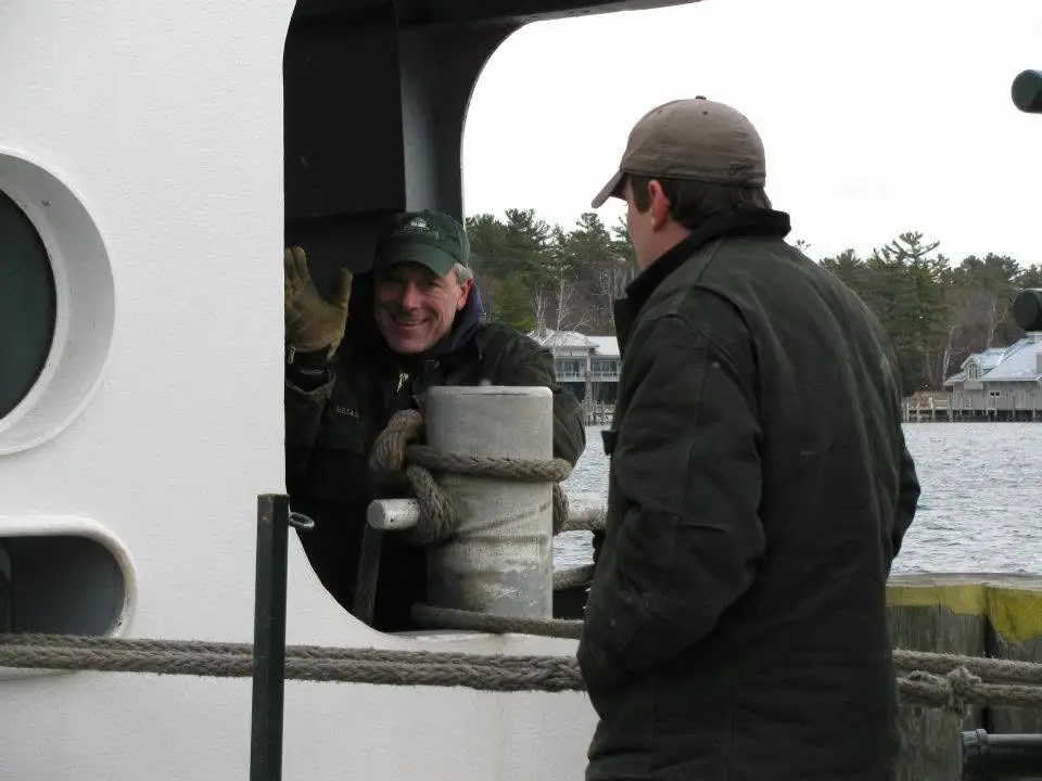 Two men stand beside a mooring bollard on a white vessel, with one waving toward the camera.