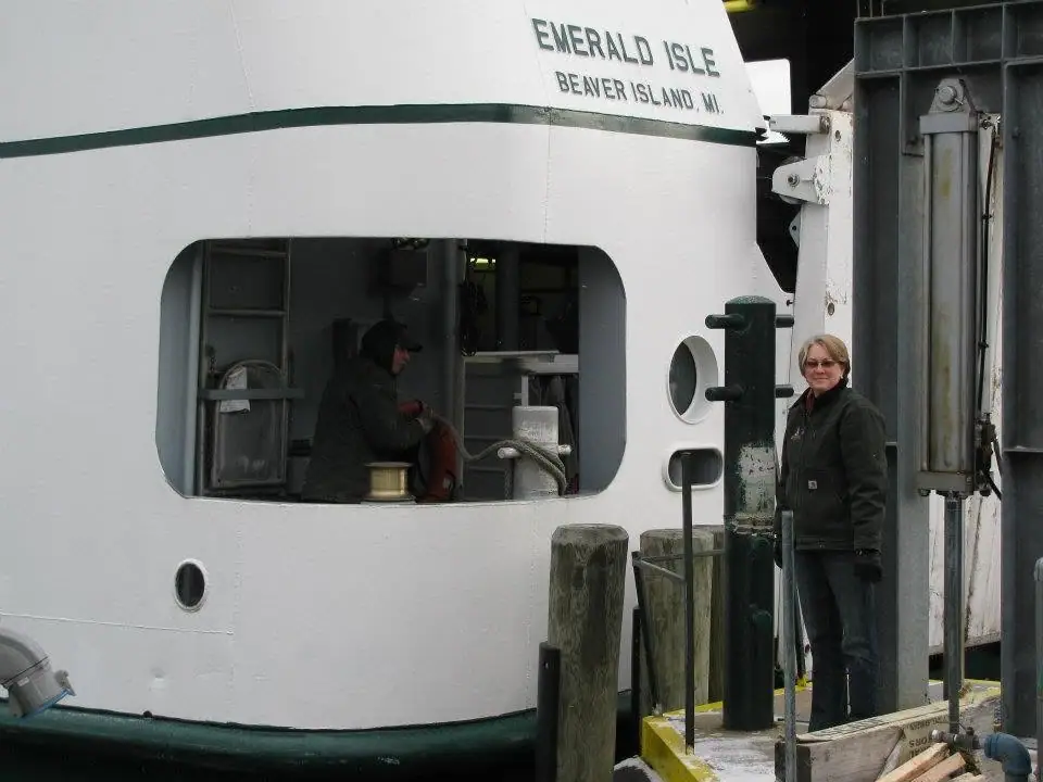 A woman stands beside a white ferry with green trim at a dock, while another person is visible through its window.