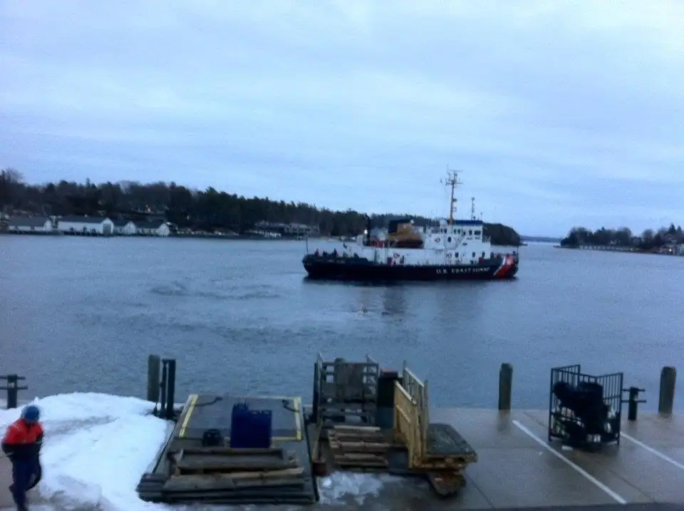 A U.S. Coast Guard cutter passes a snowy dock with loading equipment in the foreground.