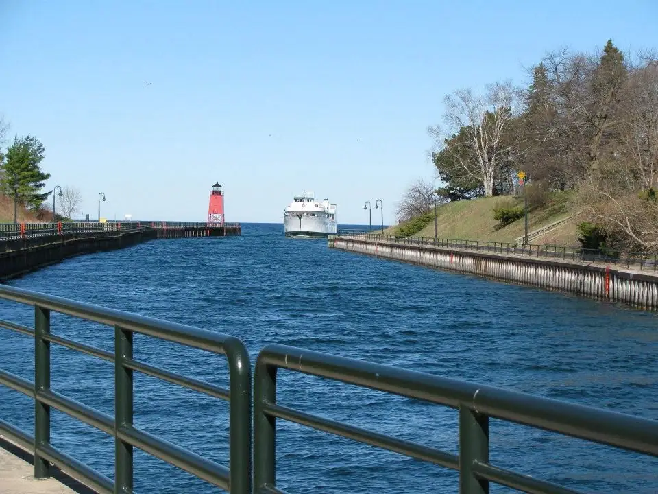 A white ferry approaches a channel beside a red lighthouse, with green railings in the foreground.