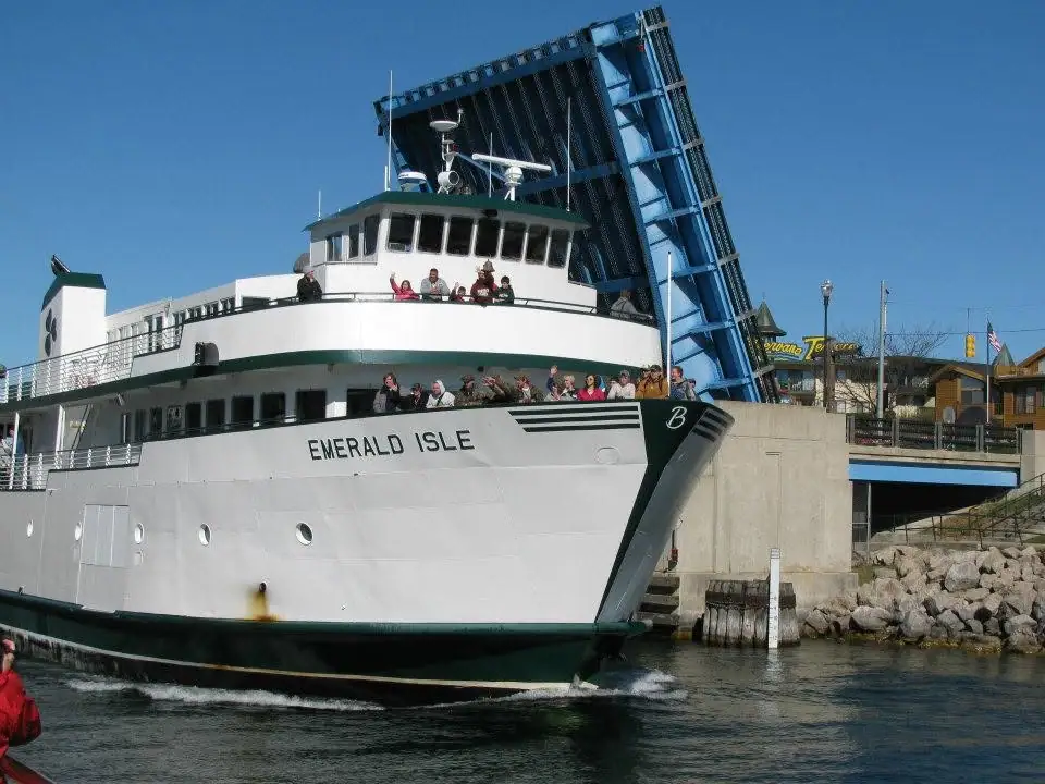 A passenger ferry carrying waving people moves beneath a raised blue drawbridge beside a waterfront.