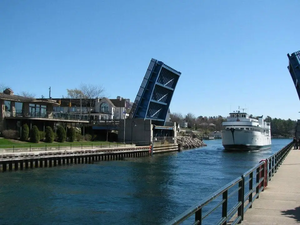 A white ferry passes beneath a raised blue drawbridge along a waterfront walkway.