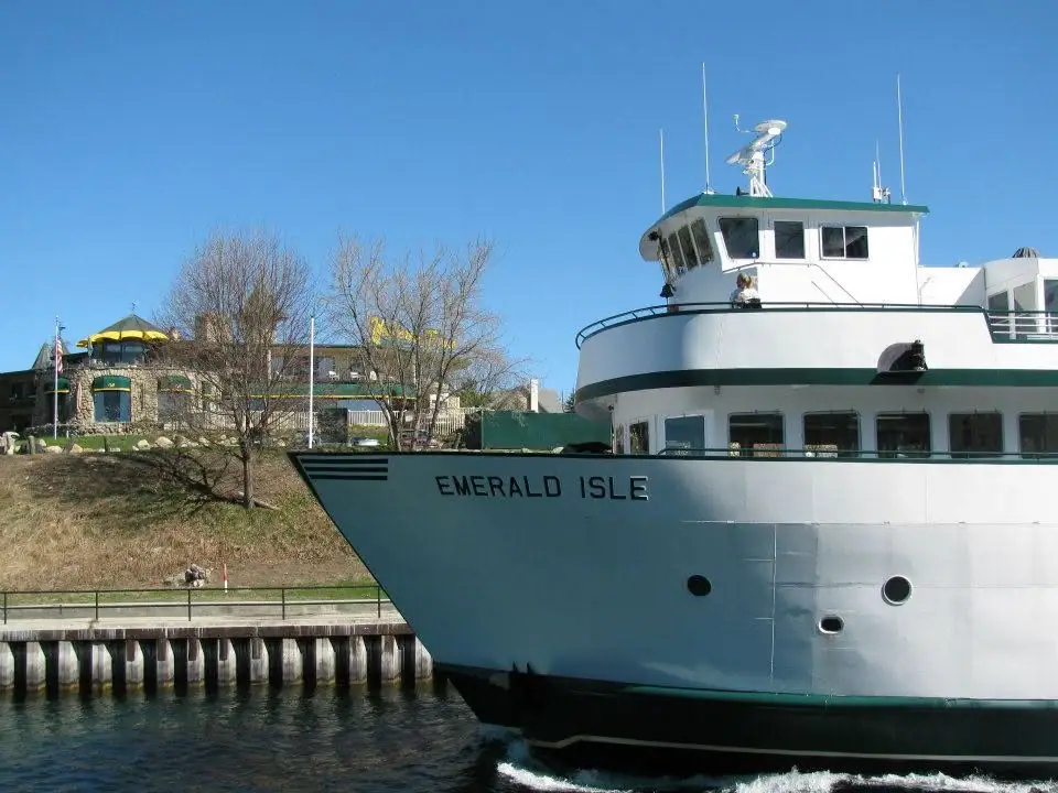 A white ferry with a dark green stripe passes a pier beside a waterfront building.