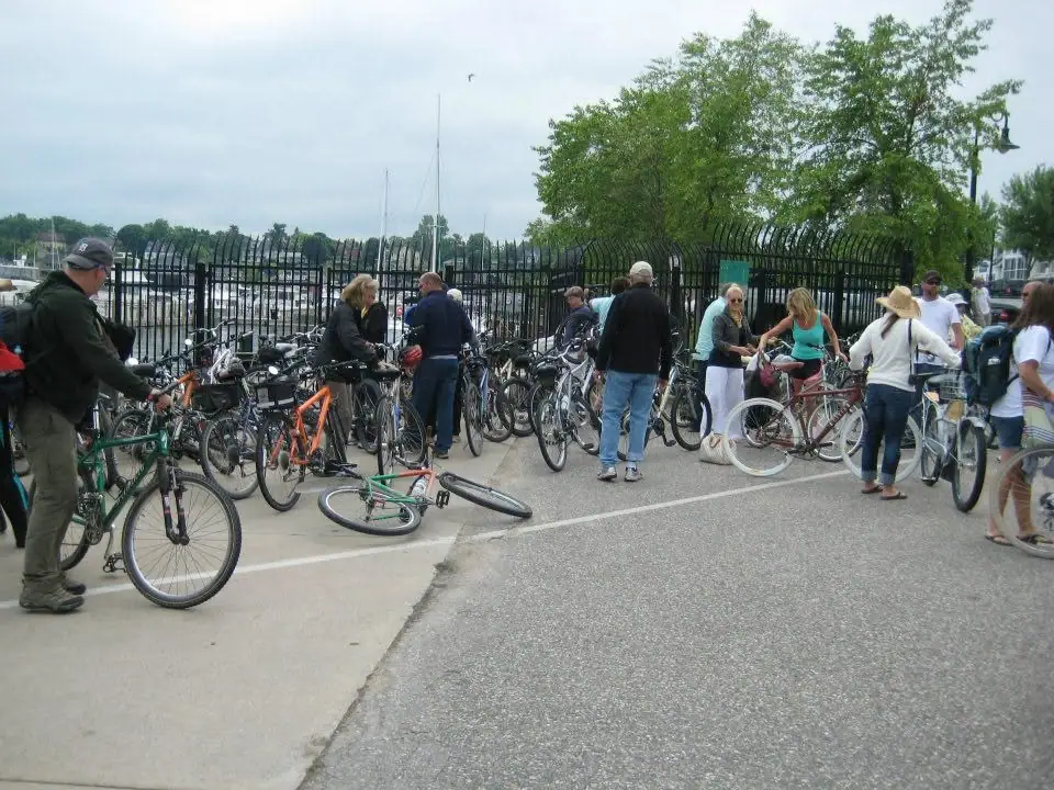 Adults gather with numerous bicycles beside a fenced waterfront, with boats and masts visible behind them.