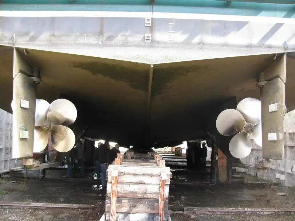 The underside of a vessel shows twin propellers, support blocks, and several people beneath the hull.