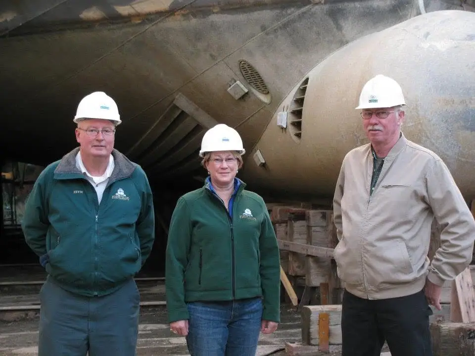 Three adults in hard hats and work jackets stand before a large weathered metal structure.