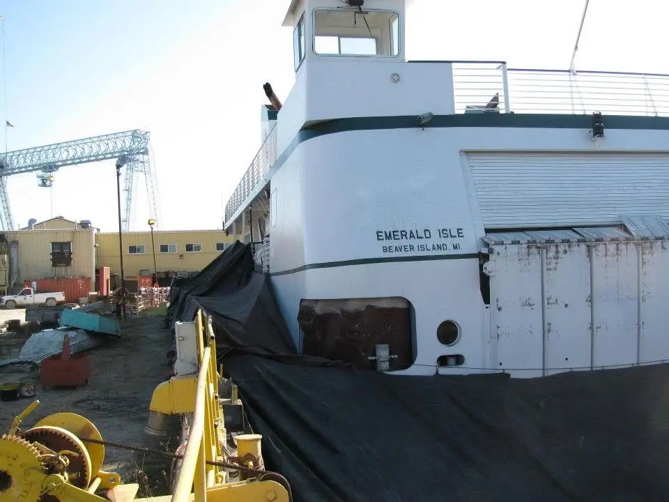 A white ferry named EMERALD ISLE stands beside industrial equipment and a crane in a maintenance yard.