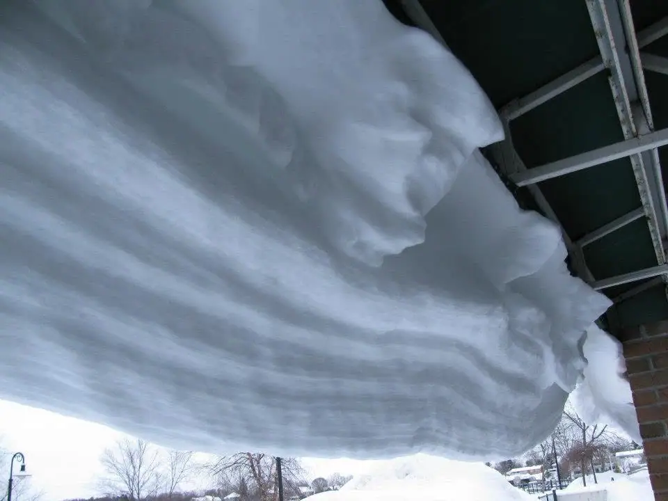 A massive bank of snow hangs from a roof over a snowy yard and leafless trees.
