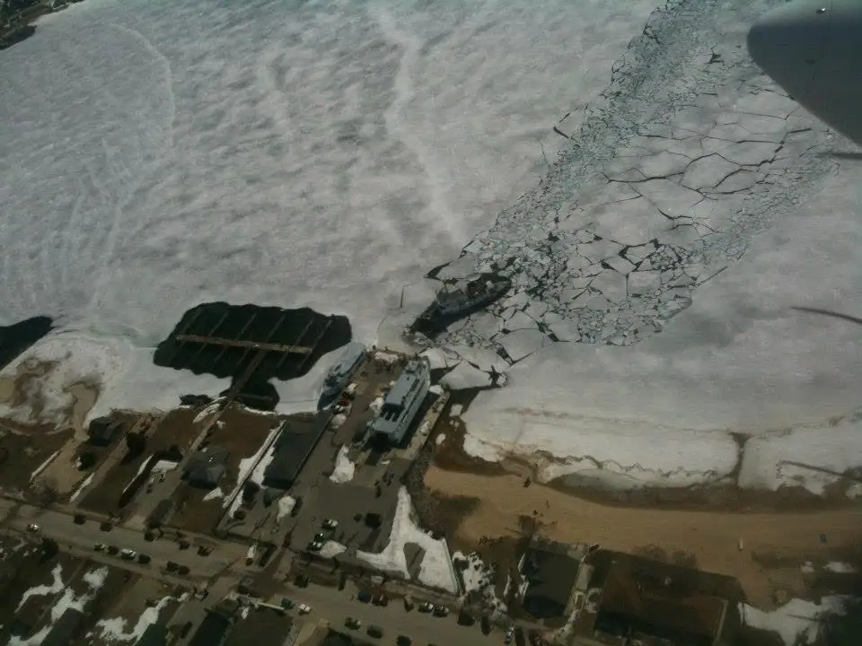 Aerial view of a ferry beside an ice-covered shoreline, with buildings, roads, and parked vehicles.