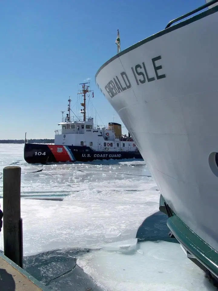 A Coast Guard cutter moves through broken ice beside the bow of a larger white vessel.