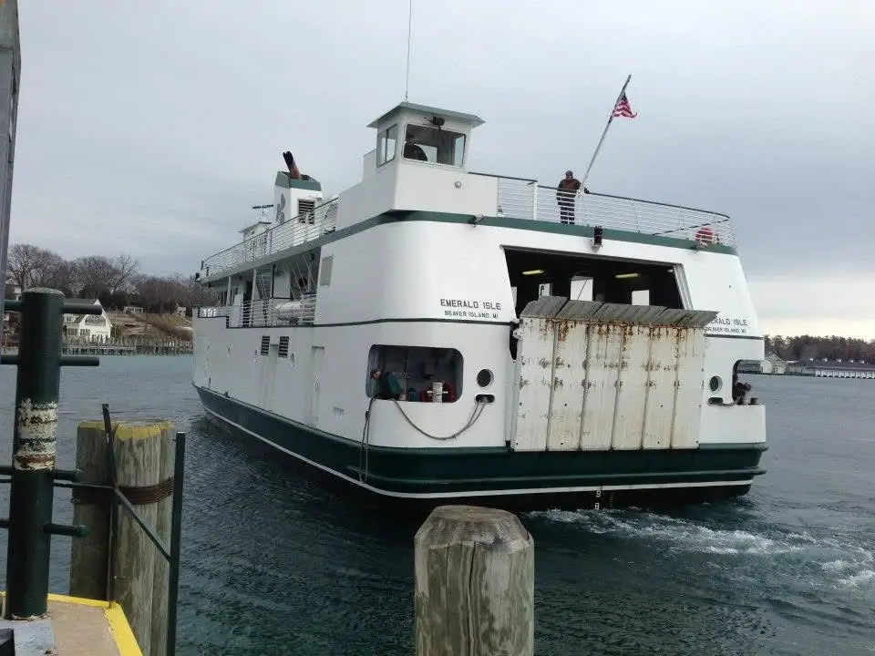 A white-and-green ferry with an open stern ramp sits beside a wooden pier, with people on deck and a flag flying.