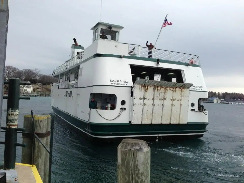 White-and-green ferry leaving a pier, with people on deck and an American flag flying above.