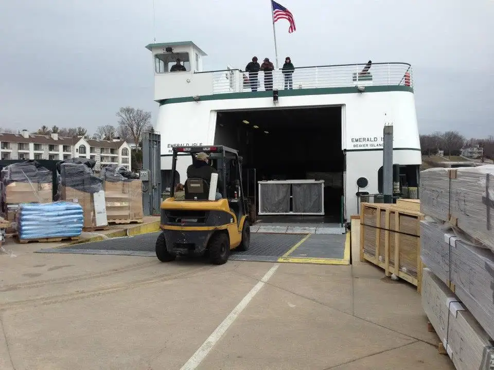 A forklift loads wooden crates into a white ferry while people stand on its upper deck.