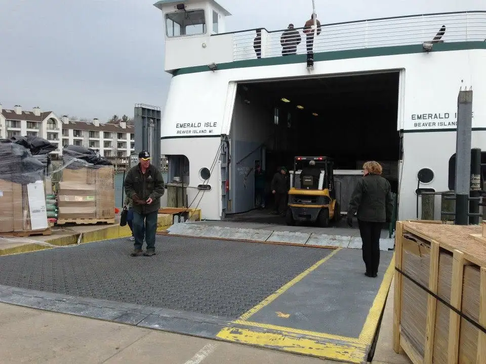 A ferry’s open loading ramp faces a dock with a forklift, cargo pallets, and several people.