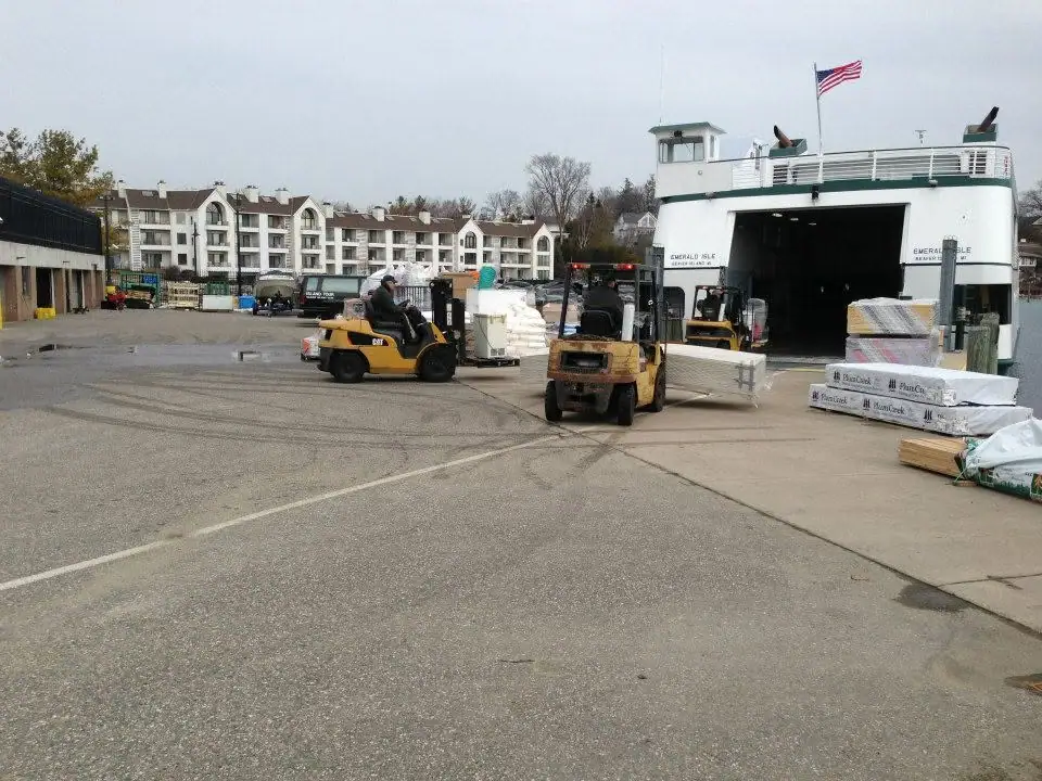 Forklifts move palletized building materials beside a ferry at a loading dock.