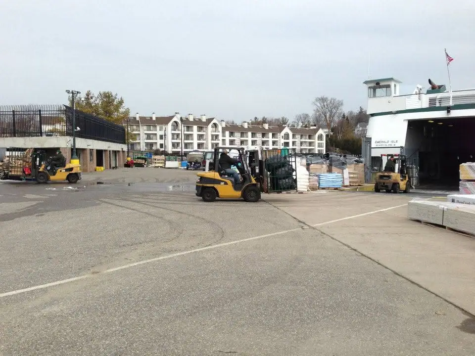 A white ferry is loaded beside three yellow forklifts and stacks of palletized goods.