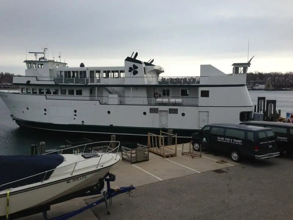 A large white ferry is moored beside a dock, with a motorboat on a trailer and two vans in the foreground.