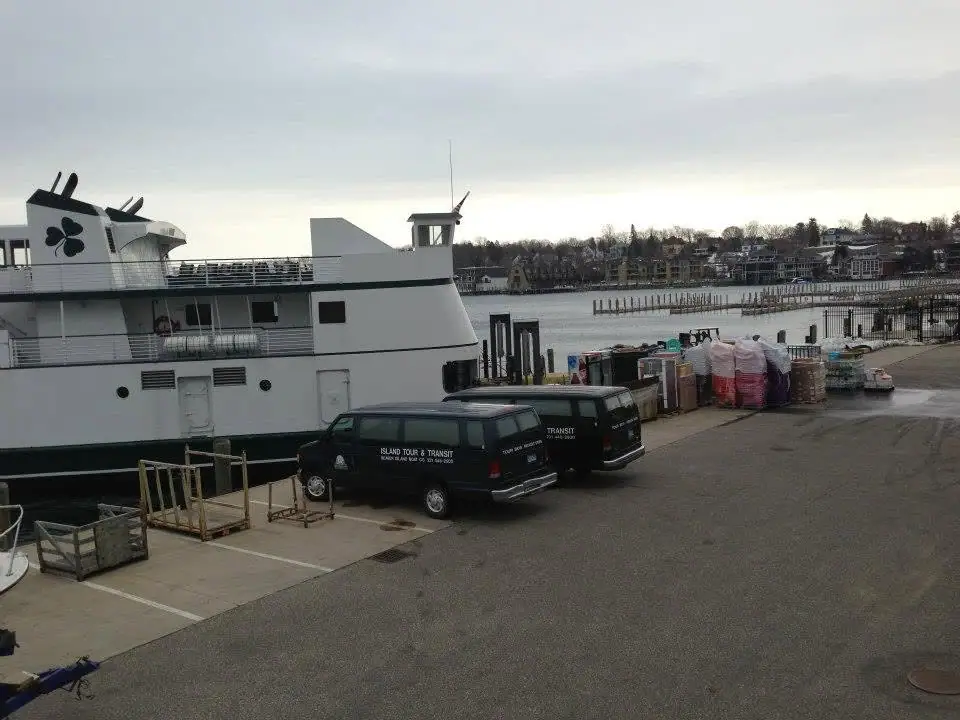 A white ferry with a shamrock emblem stands beside two vans and cargo on a waterfront dock.