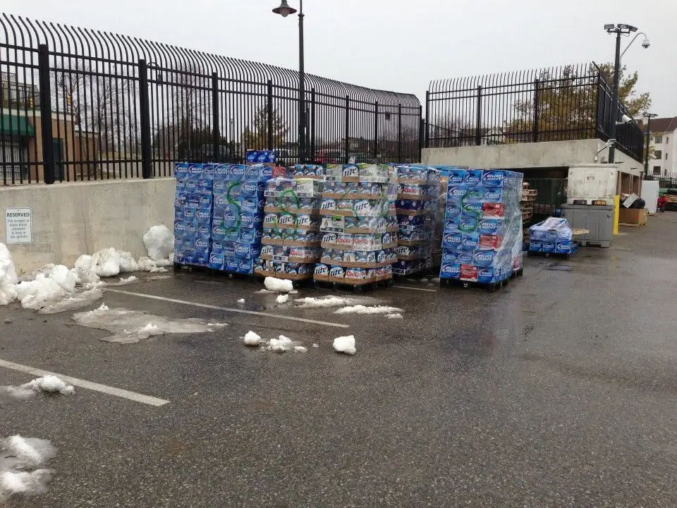 Shrink-wrapped pallets of beverage cases stand in a fenced paved lot beside melting snow.