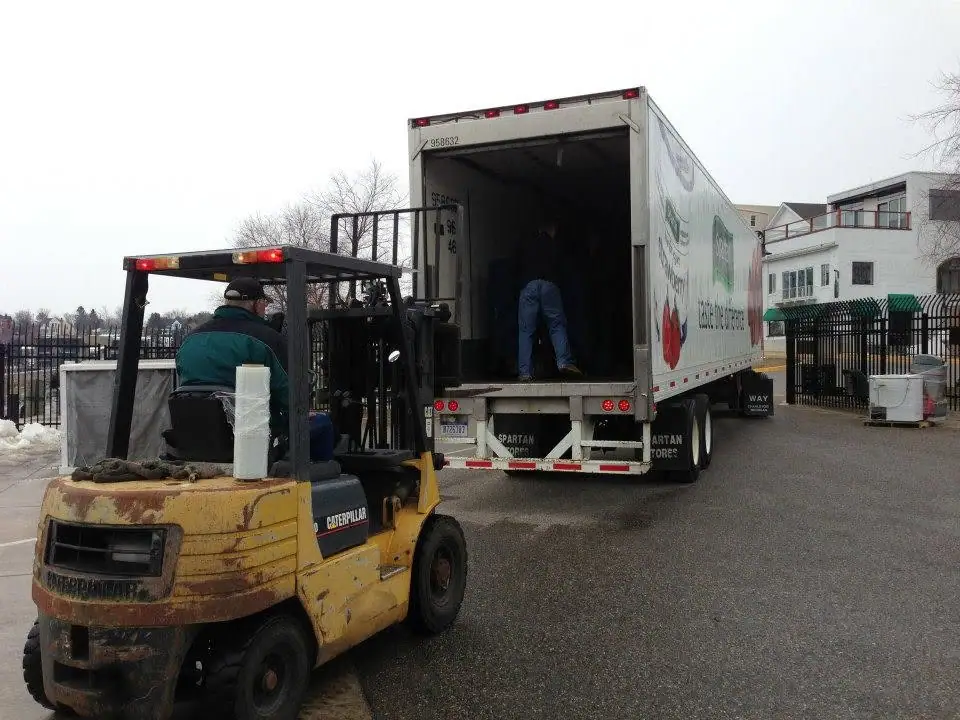 A forklift and two workers handle cargo at the open rear of a box truck in a snowy paved yard.