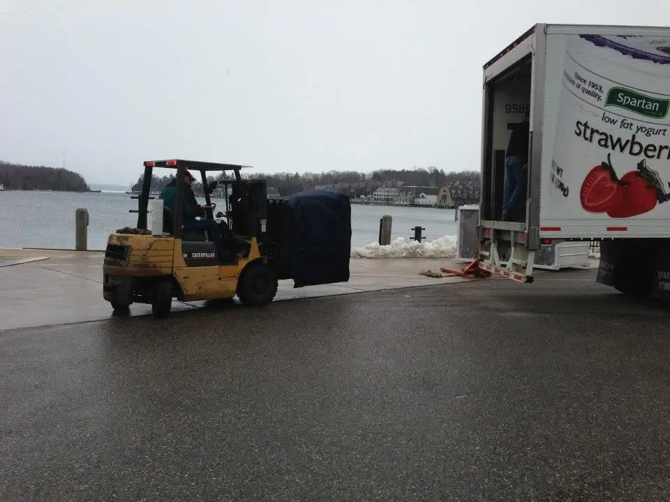 A forklift carries a covered load toward an open refrigerated delivery truck beside a waterfront dock.