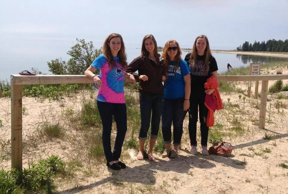 Four women pose beside a wooden railing overlooking a sandy shoreline and calm water.