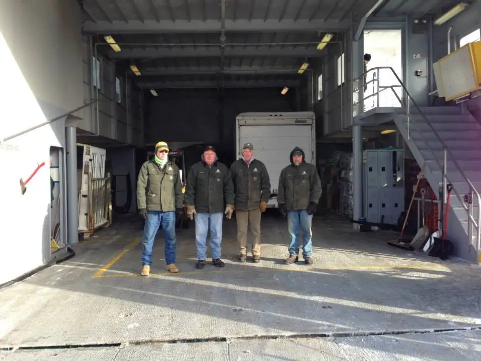 Four workers stand on an enclosed vehicle deck beside a box truck and shipboard equipment.