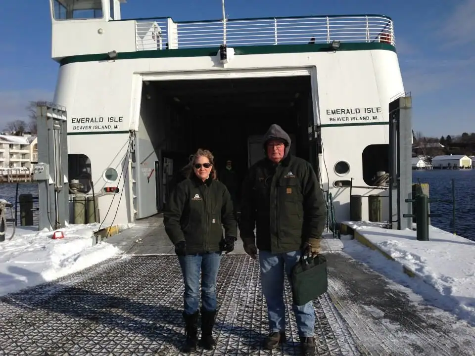 Two people stand on a snowy loading ramp in front of a white-and-green ferry with an open vehicle bay.