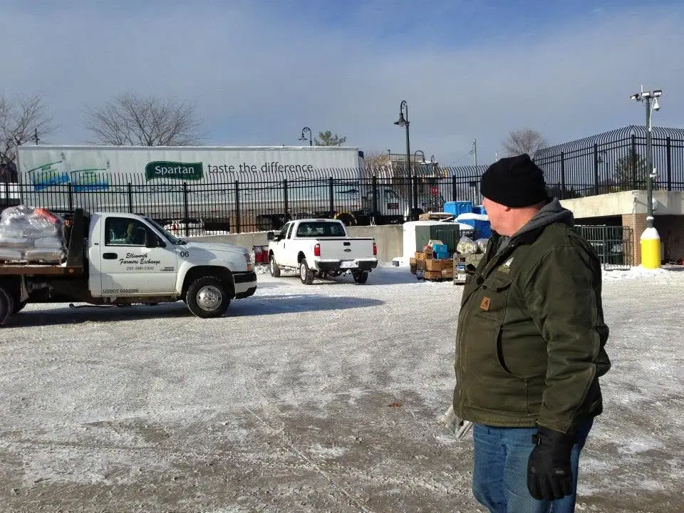 A man stands beside delivery trucks and a large trailer in a snow-covered parking area.