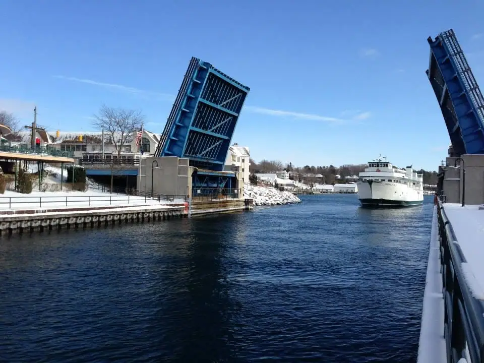 A white ferry passes between the raised spans of a blue drawbridge on a snowy waterfront.