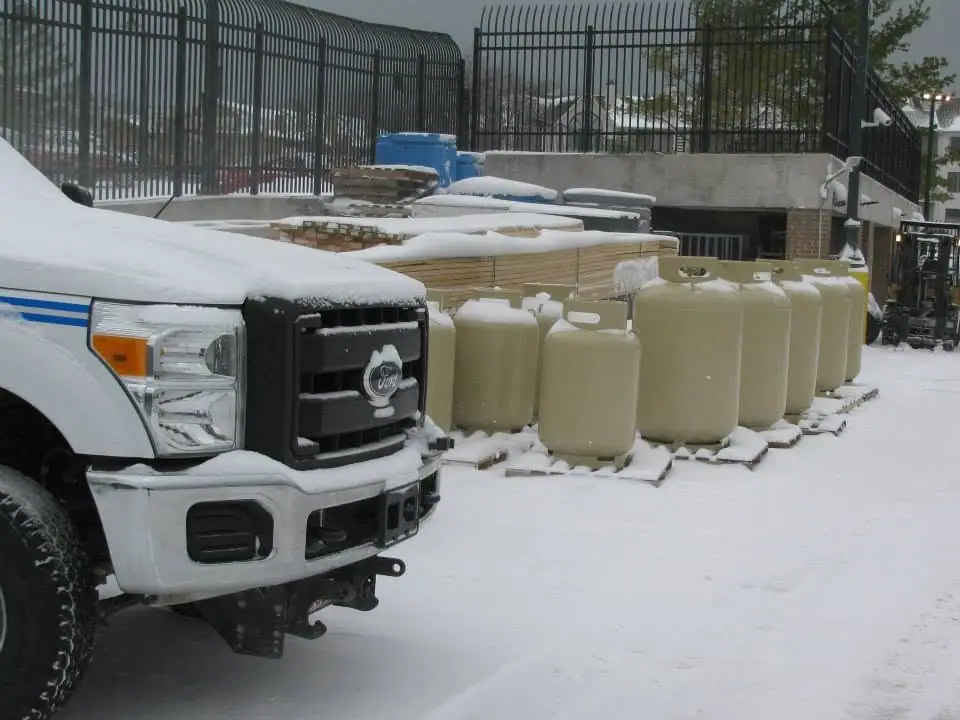 Snow-covered pickup truck beside lumber and propane tanks in a fenced outdoor yard.