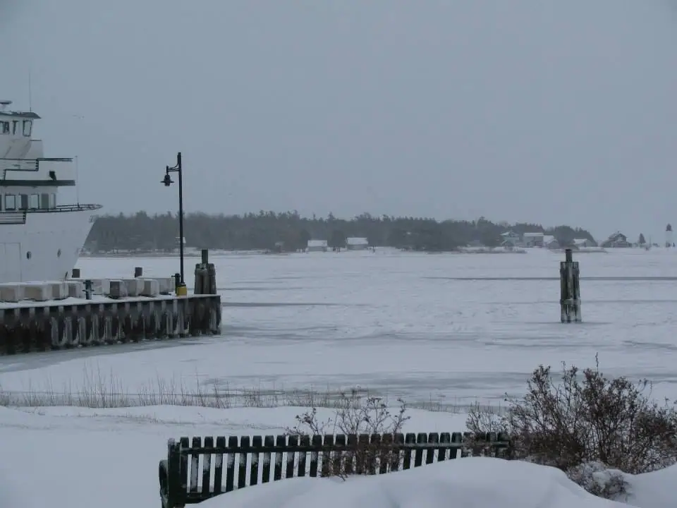 Snow-covered ferry dock and bench beside ice-covered water and a distant wooded shoreline.