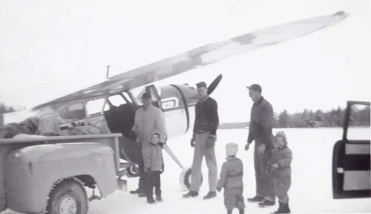 Several adults and children stand beside a small propeller airplane and pickup truck on a snow-covered field.