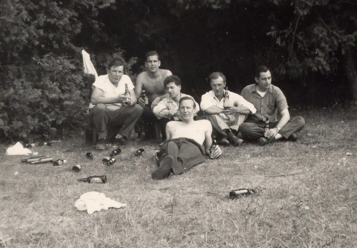 Six men sit and recline on grass in a wooded clearing, with bottles scattered around them.