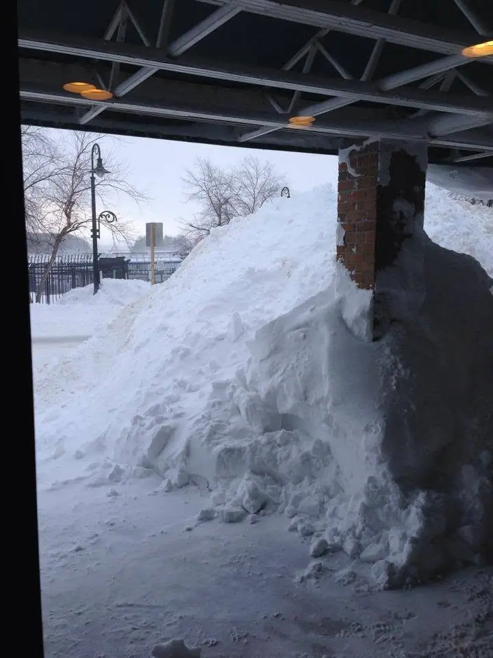 A towering snowbank beneath a steel-truss structure beside a brick pillar and decorative streetlamp.