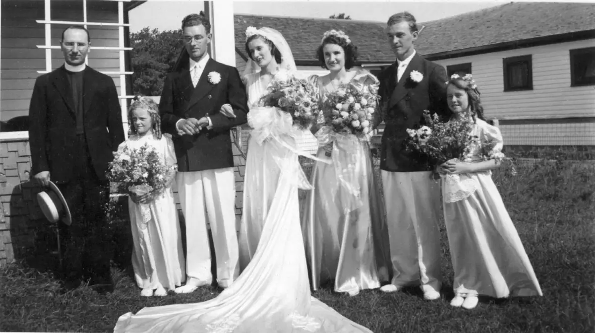 A bride, attendants, and a clergyman pose with bouquets outside a house.