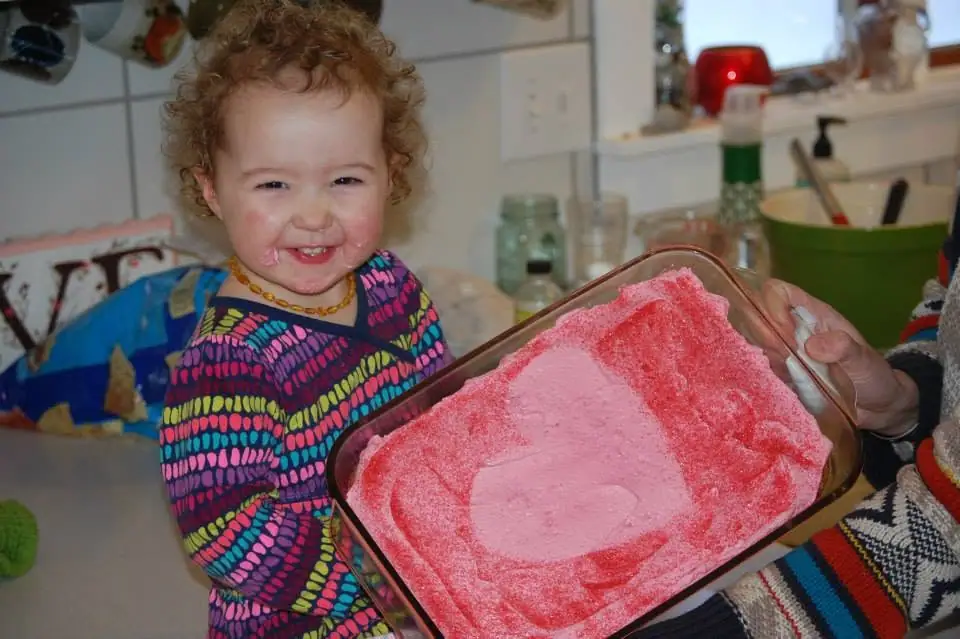 Smiling child beside a glass tray containing pink sand art shaped like a heart.