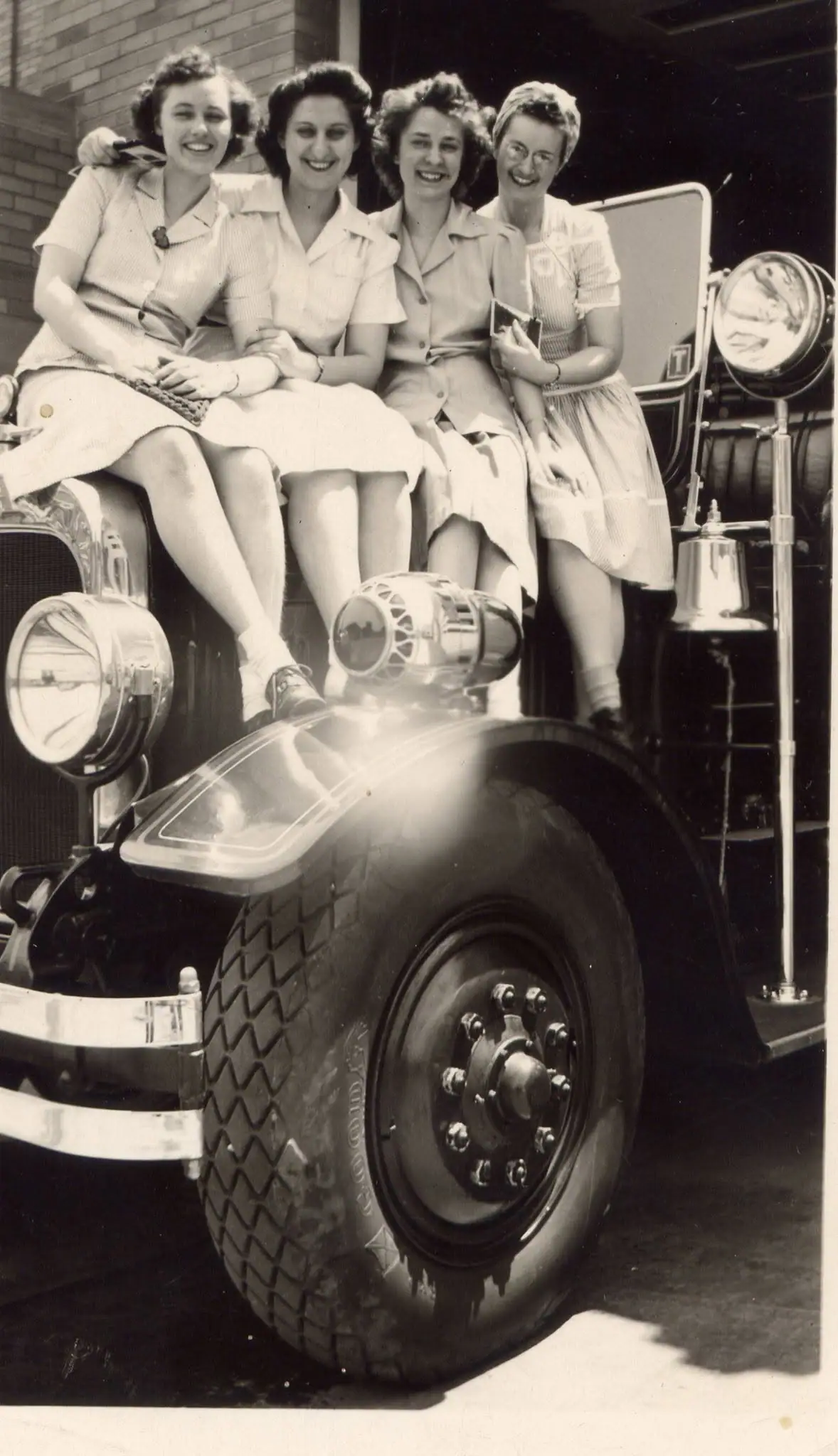 Four smiling women sit on the front of a large automobile in a black-and-white photograph.