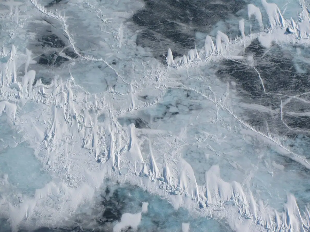 Aerial view of pale blue ice with snow-covered ridges, cracks, and dark openings.