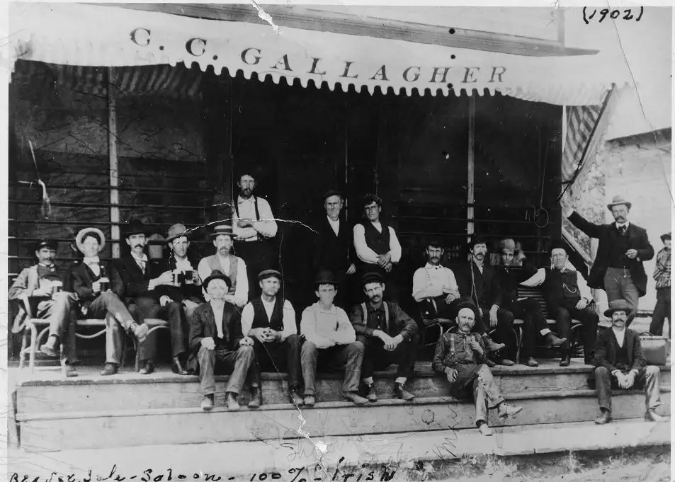 A large group of men poses on the wooden porch of a saloon beneath a C. C. Gallagher awning.