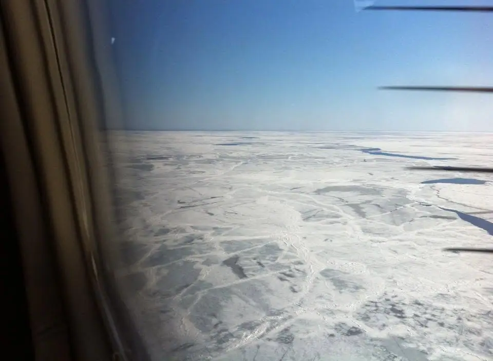 Aerial view through an airplane window of a broad snow-covered, cracked frozen surface beneath a clear sky.
