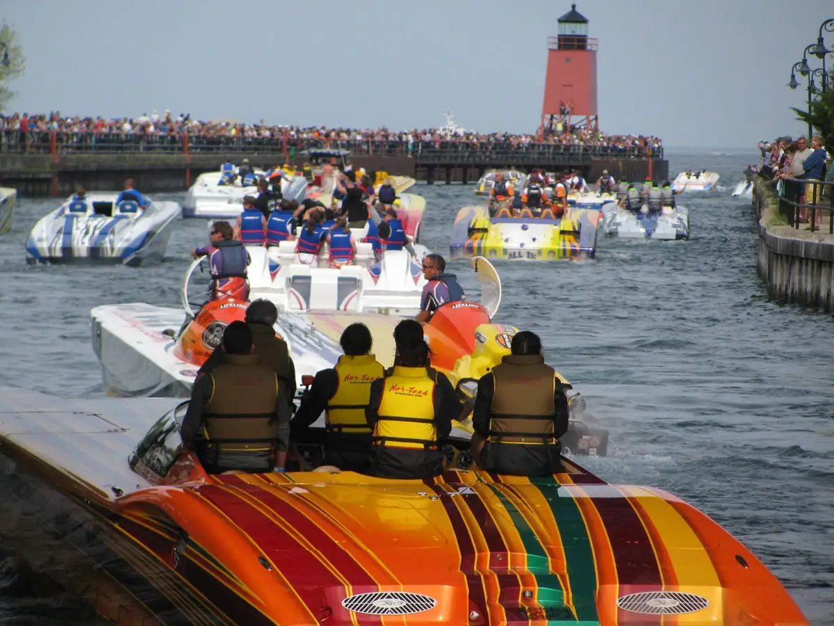 Powerboats race through a crowded harbor past a pier and red lighthouse.
