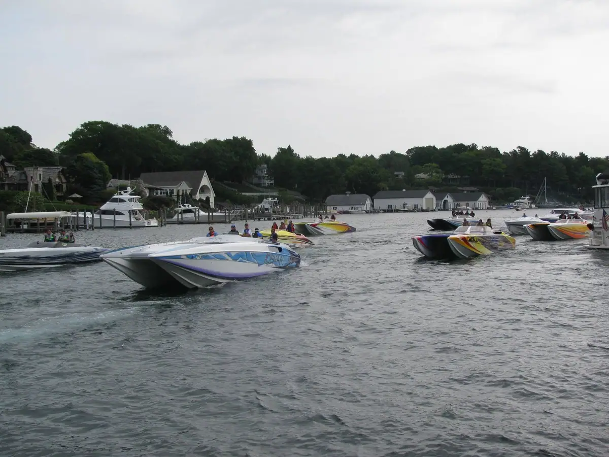 A group of colorful speedboats gathers on the water beside a wooded shoreline and marina.