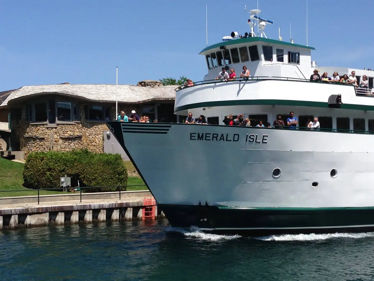 A white passenger ferry marked “EMERALD ISLE” passes a stone building and dock with people aboard.