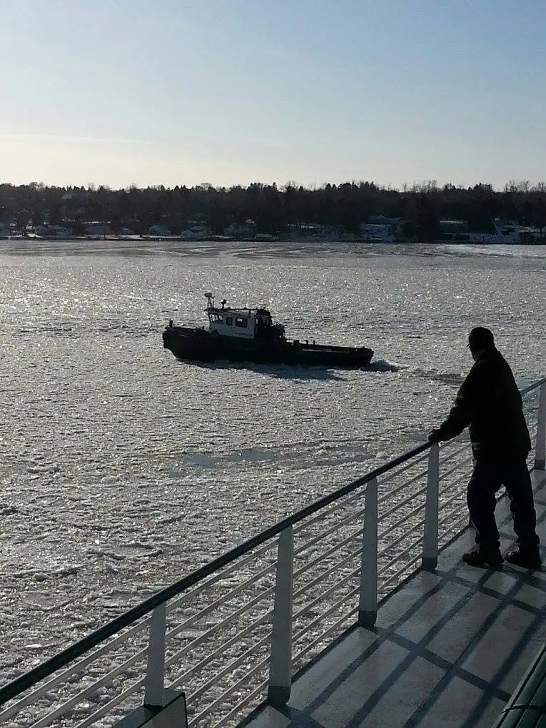 A person watches a dark workboat move through broken ice from a deck with white railings.
