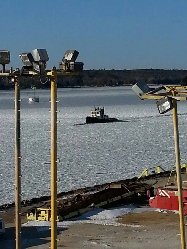 A small tugboat moves through ice beyond dockside conveyor equipment and floodlights.