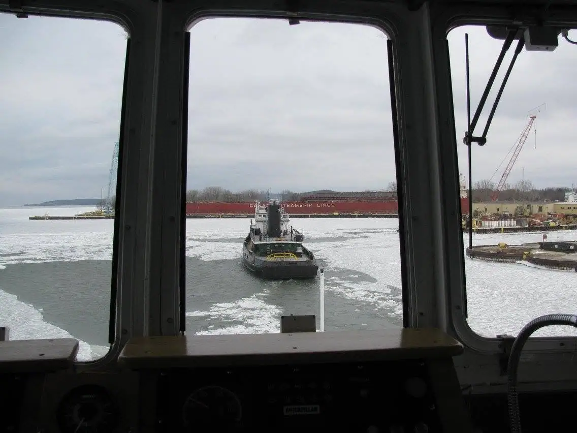 View through a vessel's bridge windows toward a tugboat, freighter, dock, crane, and ice-covered water.