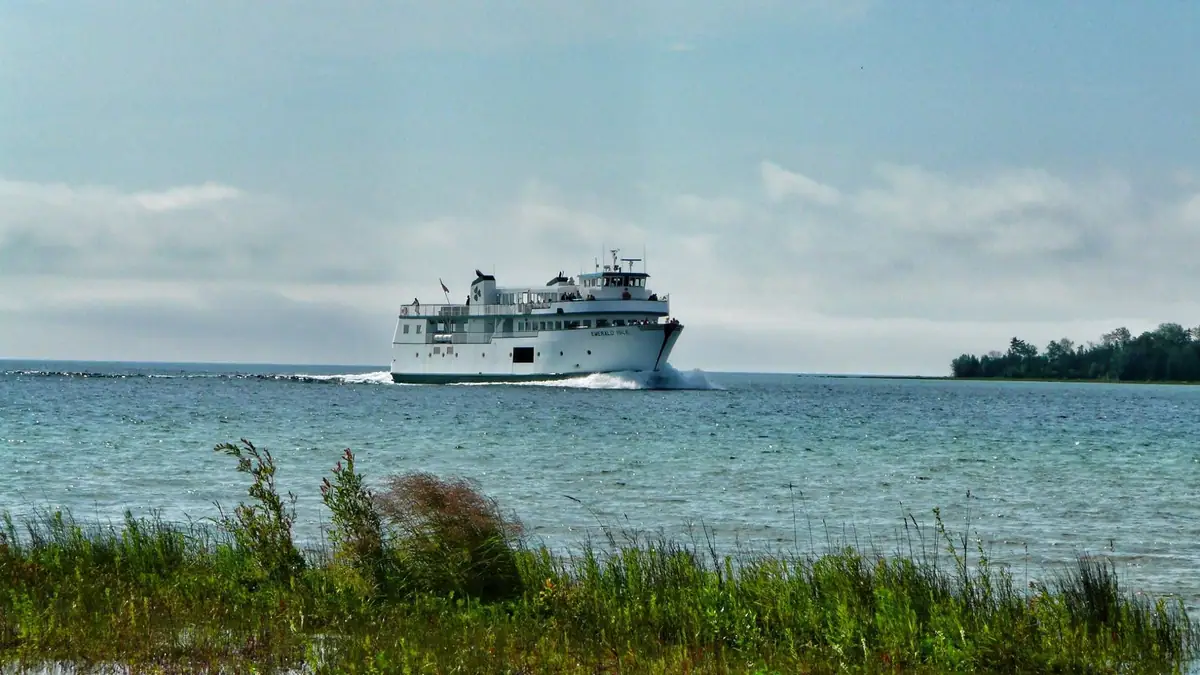 A white ferry travels across choppy water beyond a grassy shoreline, with a wooded shore in the distance.