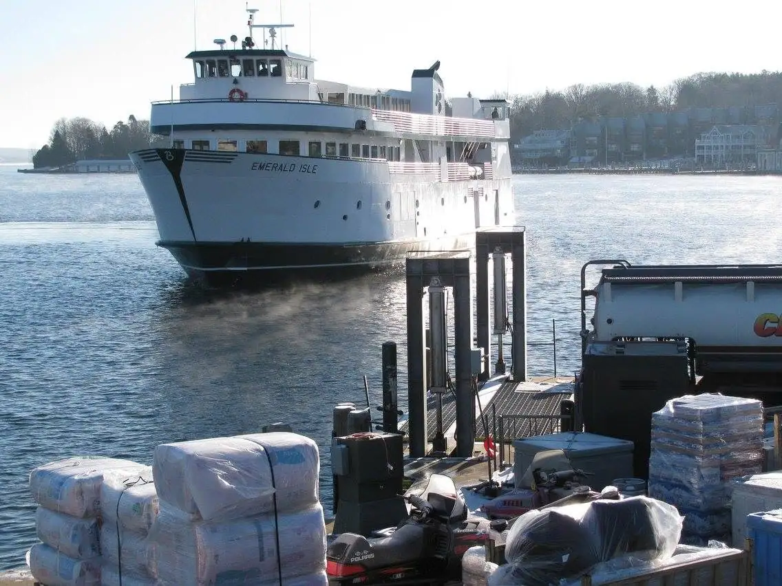 A white passenger ferry approaches a dock beside wrapped cargo, a snowmobile, and loading equipment.