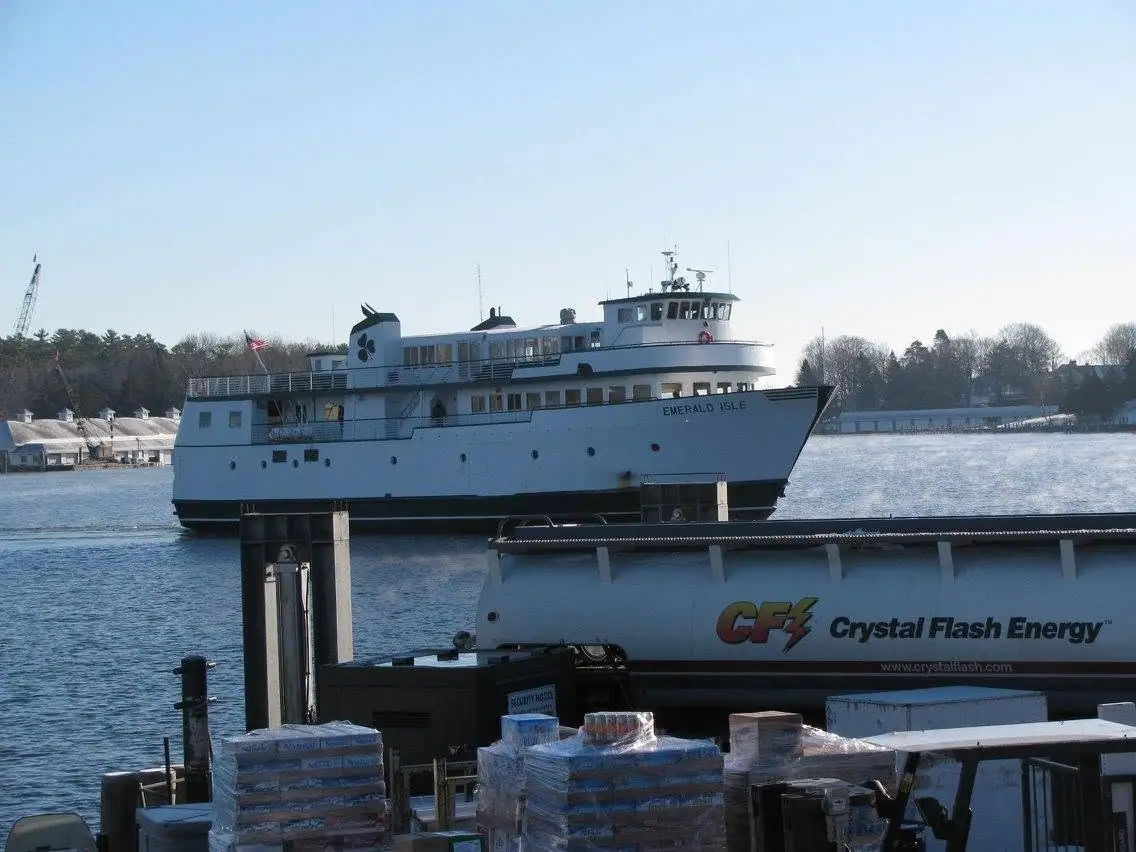 A white ferry beside a pier with a Crystal Flash Energy tanker truck and wrapped pallets in the foreground.